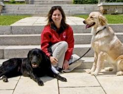 Angela, sitting on some outdoor steps, is smiling at the camera. She has a black lab lying to her left and a golden retriever sitting to her right.